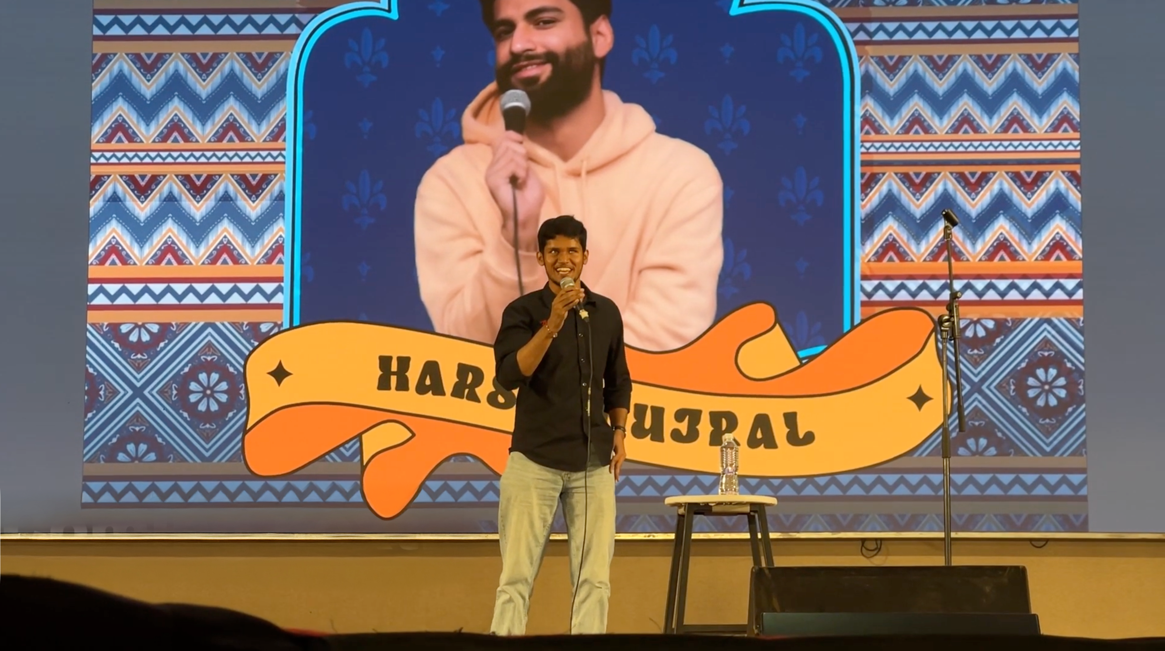 Bhavya is performing on stage with a large screen behind him showing an image of Harsh, wearing a peach hoodie, mic in hand, and his name written in bold black letters